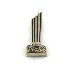 Op de afbeelding: Een zilverkleurige metalen broche in de vorm van een afgebroken klassieke zuil. Het object heeft een rechthoekige voet, een kapiteel met twee spiraalvormige voluten en een verticale schacht met vier duidelijke cannelures in de lengte.