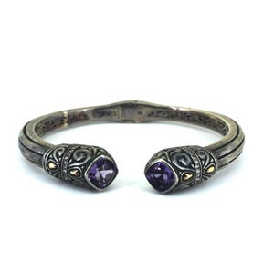 Può includere: Bracciale a polsino color argento con dettagli ornati e due pietre preziose viola quadrate. Il bracciale presenta dettagli dorati e un motivo decorativo. Un elegante gioiello.