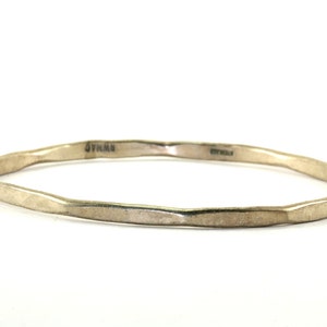 May include: A silver-toned bangle bracelet with a faceted, irregular design. The bracelet has a hammered texture and is marked with the word "STERLING". The overall style is minimalist and modern.