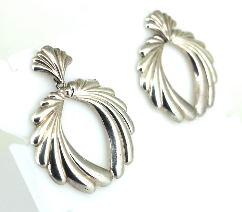 May include: A pair of silver dangle earrings with a fan-shaped design. The earrings are made of smooth, polished metal and have a modern, geometric look.