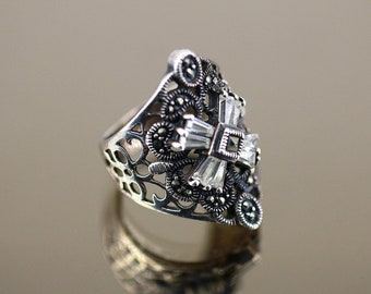 May include: A silver ring with a filigree design and a cross-shaped center. The cross is adorned with clear cubic zirconia stones and black marcasite stones.