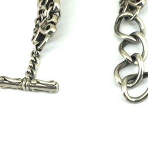 May include: Close-up of a silver-toned bracelet with a toggle clasp. The bracelet features a chain design with a series of interconnected links. The toggle clasp is a bar shape. The jewelry is set against a white background.