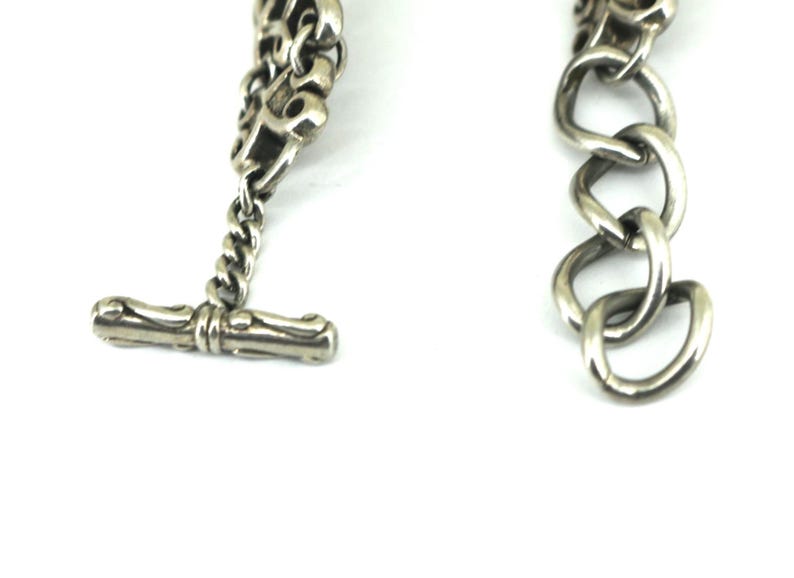 May include: Close-up of a silver-toned bracelet with a toggle clasp. The bracelet features a chain design with a series of interconnected links. The toggle clasp is a bar shape. The jewelry is set against a white background.