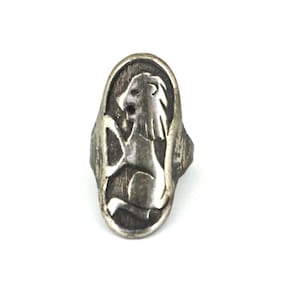 May include: An oval-shaped silver-toned ring featuring a raised, stylized lion figure in profile. The lion is depicted in a rampant position against a textured, dark background within the oval frame, showcasing a metallic finish and detailed relief.