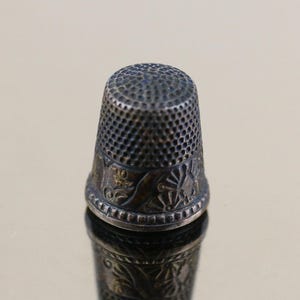 Op de afbeelding: Een vintage zilveren vingerhoed met een gedetailleerd, getextureerd oppervlak. De vingerhoed heeft een decoratieve band met bloemen en geometrische patronen. De bovenkant is ingedeukt voor naaldgrip.