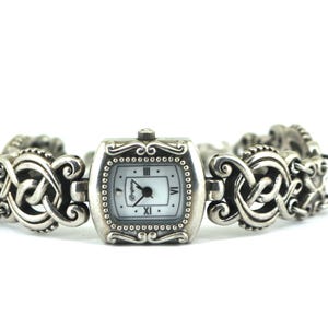 May include: A silver-toned watch with a rectangular face and a decorative band. The watch face is white with black hands and Roman numeral hour markers. The band features an intricate, Celtic-inspired design.