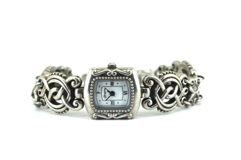 May include: A silver-toned watch with a rectangular face and a decorative band. The watch face is white with black hands and Roman numeral hour markers. The band features an intricate, Celtic-inspired design.