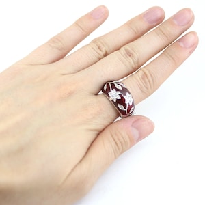 May include: A maroon ring with a floral design. The ring features white flowers and leaves set against a maroon background. The ring is on a finger.