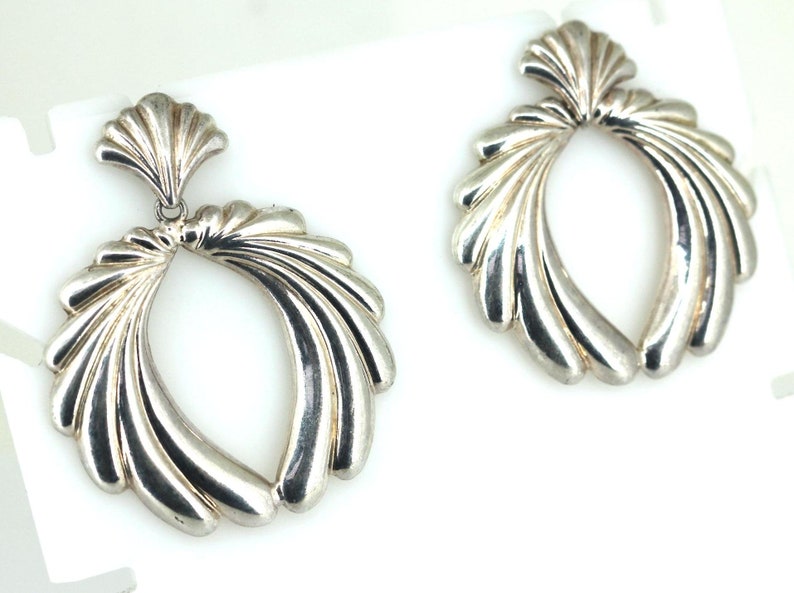 May include: A pair of silver earrings with a fan-shaped design. The earrings are made of metal and have a shiny finish.