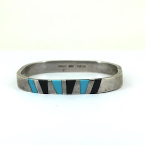 Peut inclure: Un bracelet argenté de forme rectangulaire. Le bracelet présente un motif d'incrustations triangulaires noires et turquoise alternées. Le bracelet est présenté sur une surface blanche.
