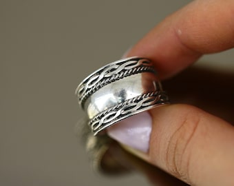 May include: A silver ring with a wide band and a braided design. The band is smooth and polished.