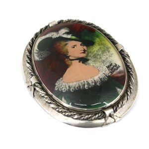 May include: A silver oval brooch with a painted portrait of a woman wearing a green dress and a white hat. The painting is signed "After Charles Bertrand".