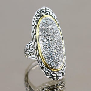 May include: A silver and gold ring with a large oval-shaped center stone set with small clear stones. The ring has a detailed design with a braided pattern.