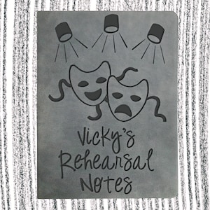 May include: A gray leather notebook with a black design of two theater masks and spotlights. The text "Vicky's Rehearsal Notes" is printed on the front.