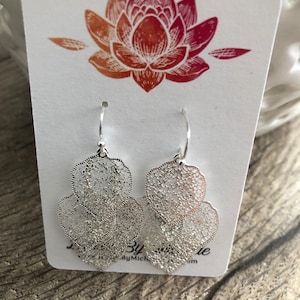 May include: Silver filigree earrings with a delicate, openwork design. The earrings are displayed on a white card with a lotus flower illustration in shades of red and orange. The earrings are on a wooden surface.