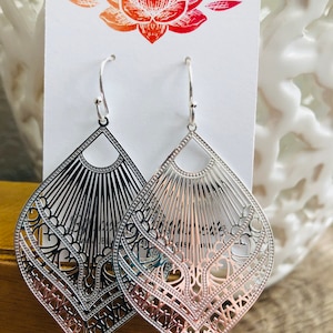 May include: A pair of silver filigree earrings with a teardrop shape and intricate designs. The earrings are hanging from a white card with a red and orange lotus flower design.