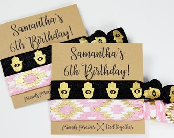 May include: A collection of elastic hair ties with accompanying cards. The cards display the text "Samantha's 6th Birthday!" and "friends forever tied together." The hair ties feature black bands with gold hamsa hands and pink and white patterns.