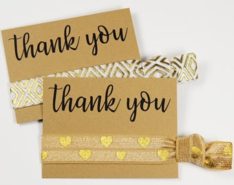 May include: Two brown cards with the text "thank you" written in black cursive. The cards are tied together with two gold and white patterned hair ties.