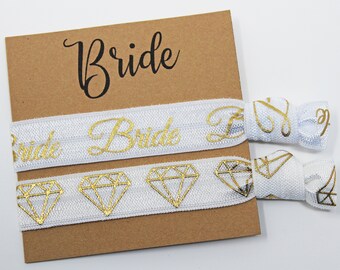 May include: Two white elastic hair ties with gold foil lettering. One hair tie says "Bride" and the other has a diamond pattern. The hair ties are on a brown card with the word "Bride" written in black.