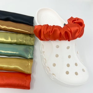 May include: A white clog shoe with an orange satin scrunchie attached. Several satin scrunchies in various colors, including black, gold, green, and orange, are stacked to the left of the shoe. The scrunchies appear to be accessories for the clog.