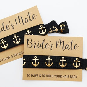 Może przedstawiać: Dwa czarne gumki do włosów z złotymi wzorami kotwic i brązowa karta z napisem "Bride's Mate" i "To Have & To Hold Your Hair Back".