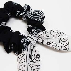 Op de afbeelding: Een zwart-wit bandana haarband met een paisley-patroon. De haarband heeft twee lange staarten die aan elkaar zijn geknoopt.