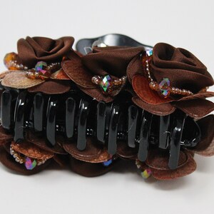 Może przedstawiać: Brązowa spinka do włos&oacute;w z dużym wzorem kwiatowym. Kwiat wykonany jest z brązowej tkaniny i ozdobiony jest tęczowymi koralikami i kryształkami.