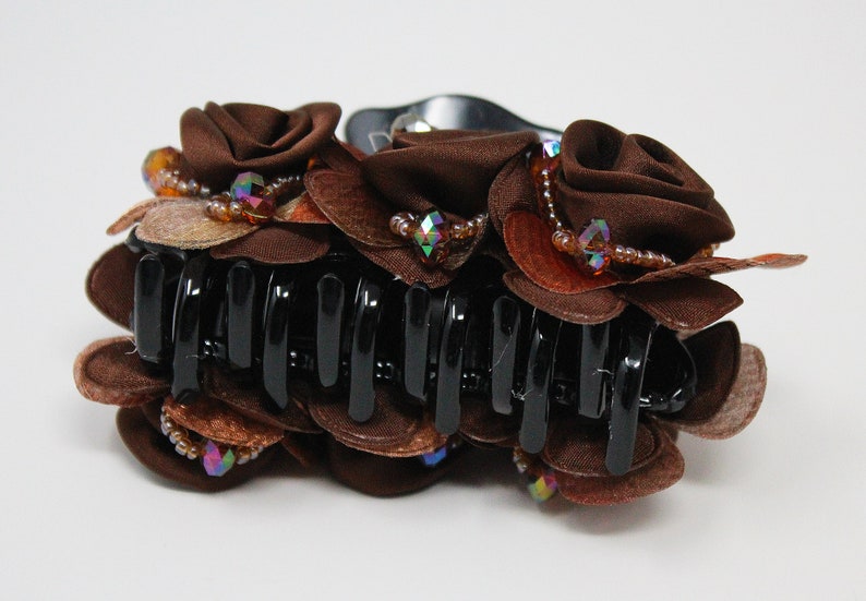 Może przedstawiać: Brązowa spinka do włos&oacute;w z dużym wzorem kwiatowym. Kwiat wykonany jest z brązowej tkaniny i ozdobiony jest tęczowymi koralikami i kryształkami.