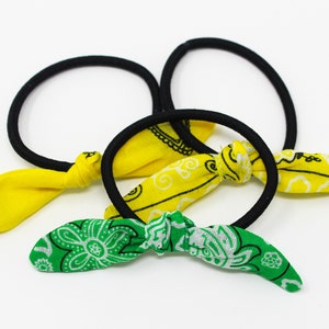 Puede incluir: Tres gomas para el pelo con lazos de tela. Dos lazos son amarillos con estampado paisley negro y un lazo es verde con estampado paisley blanco.