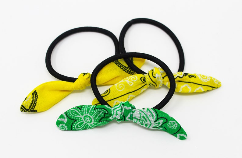 Puede incluir: Tres gomas para el pelo con lazos de tela. Dos lazos son amarillos con estampado paisley negro y un lazo es verde con estampado paisley blanco.