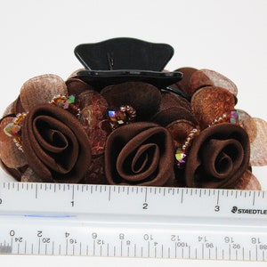 Może przedstawiać: Brązowa spinka do włos&oacute;w z czarną plastikową podstawą i brązowymi r&oacute;żami z tkaniny. Spinka jest ozdobiona tęczowymi koralikami i brązową siateczką.