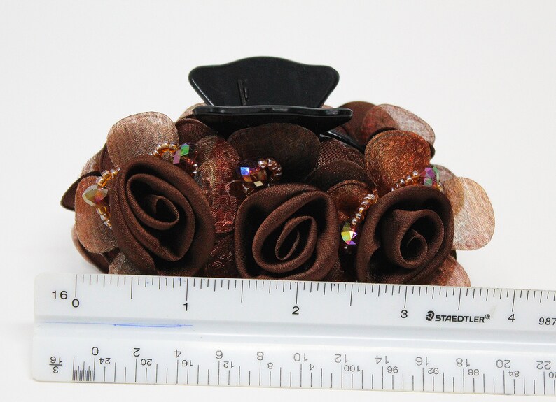 Może przedstawiać: Brązowa spinka do włos&oacute;w z czarną plastikową podstawą i brązowymi r&oacute;żami z tkaniny. Spinka jest ozdobiona tęczowymi koralikami i brązową siateczką.