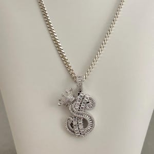 Puede incluir: Collar de cadena de plata con un colgante con una letra 'S' con una corona en la parte superior, ambos incrustados con piedras de circonita cúbica transparentes.