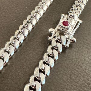 May include: A silver-toned metal cuban link chain necklace featuring a box clasp closure. The clasp is accented with a single, small, round, deep red gemstone set in the center of the rectangular locking mechanism, providing a distinct focal point.