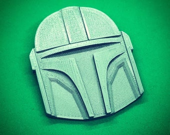 May include: A silver 3D printed Mandalorian helmet. The helmet is detailed with lines and grooves.