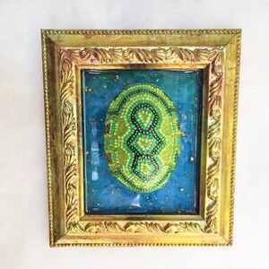 May include: A green and black beaded egg in a gold frame with a blue background. The egg is made of small beads and has a geometric pattern.