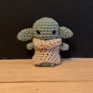 Peut inclure: Une petite poupée crochetée à la main avec une tête vert clair, de grandes oreilles pointues et deux yeux en perles noires. La figurine porte un vêtement beige texturé et ample qui recouvre son torse et ses bras, réalisé avec des mailles visibles.