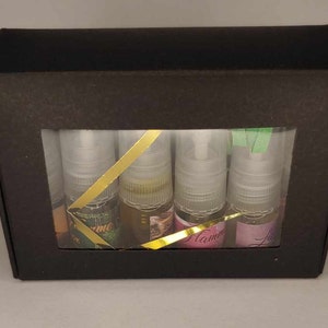 Puede incluir: Una caja de regalo rectangular negra con una ventana transparente que muestra cuatro pequeños frascos rociadores transparentes. Cada botella contiene un líquido de diferente color y tiene una tapa plateada. Cintas doradas acentúan las botellas. Las botellas tienen etiquetas con texto.