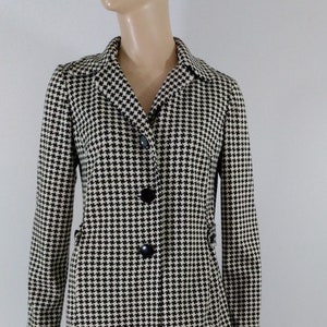 May include: A black and white houndstooth patterned coat with a long sleeve and a button closure.