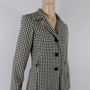 May include: A black and white houndstooth patterned coat with a long sleeve and a button closure.