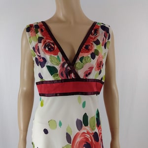 Puede incluir: Un vestido blanco sin mangas con estampado floral de rosas rosas, rojas y moradas. El vestido tiene un escote en V y una cinta de color burdeos y roja alrededor de la cintura.