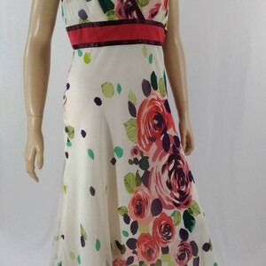 Puede incluir: Un vestido blanco sin mangas con estampado floral con rosas rojas, rosas y moradas. El vestido tiene un escote en V y una cintura roja y burdeos.