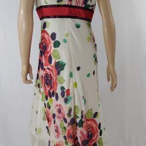 Puede incluir: Un vestido blanco sin mangas con estampado floral de rosas rosas, rojas y verdes. El vestido tiene una cintura roja y burdeos y un escote en V.