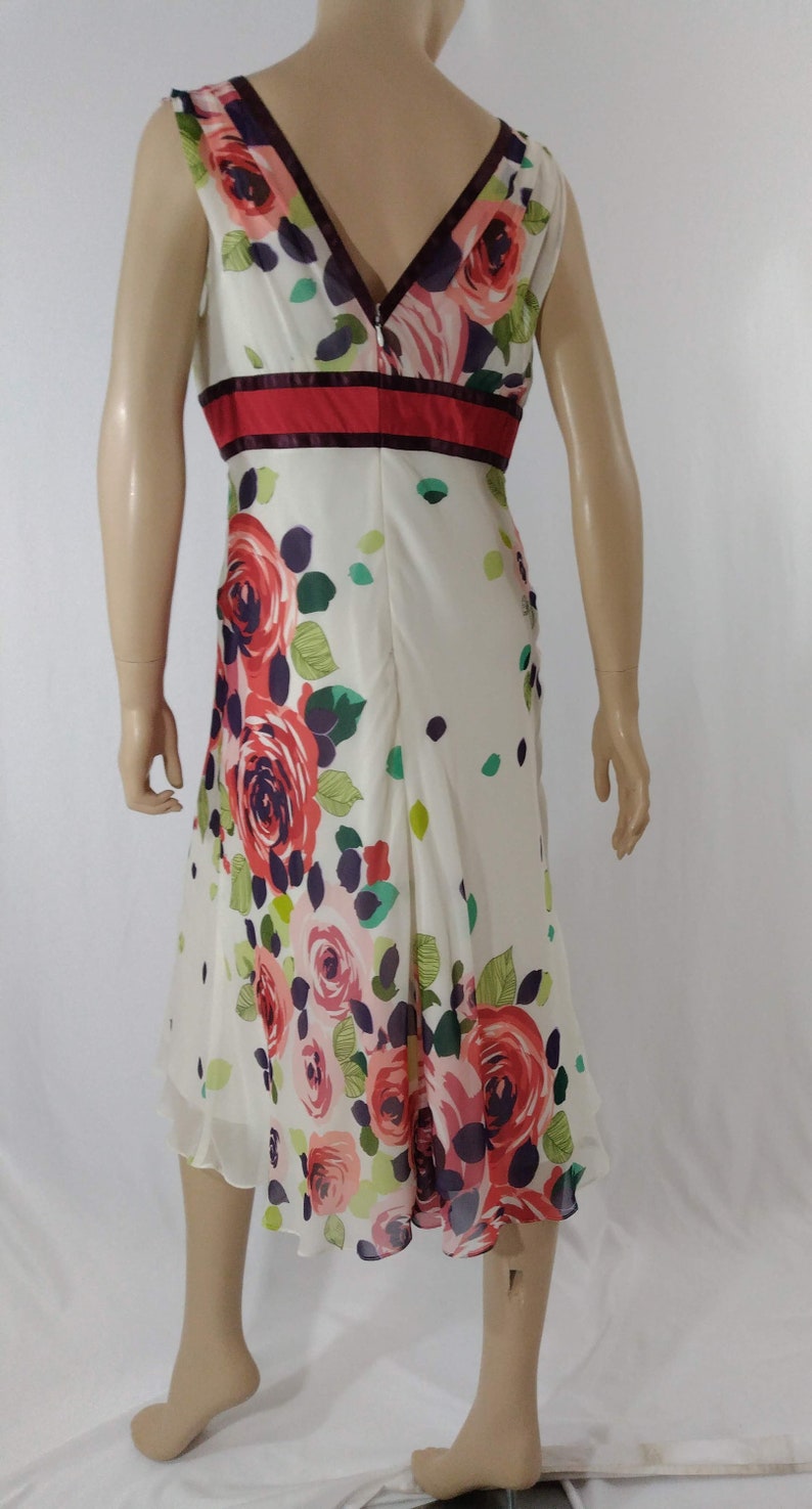 Puede incluir: Un vestido blanco sin mangas con estampado floral de rosas rosas, rojas y verdes. El vestido tiene una cintura roja y burdeos y un escote en V.