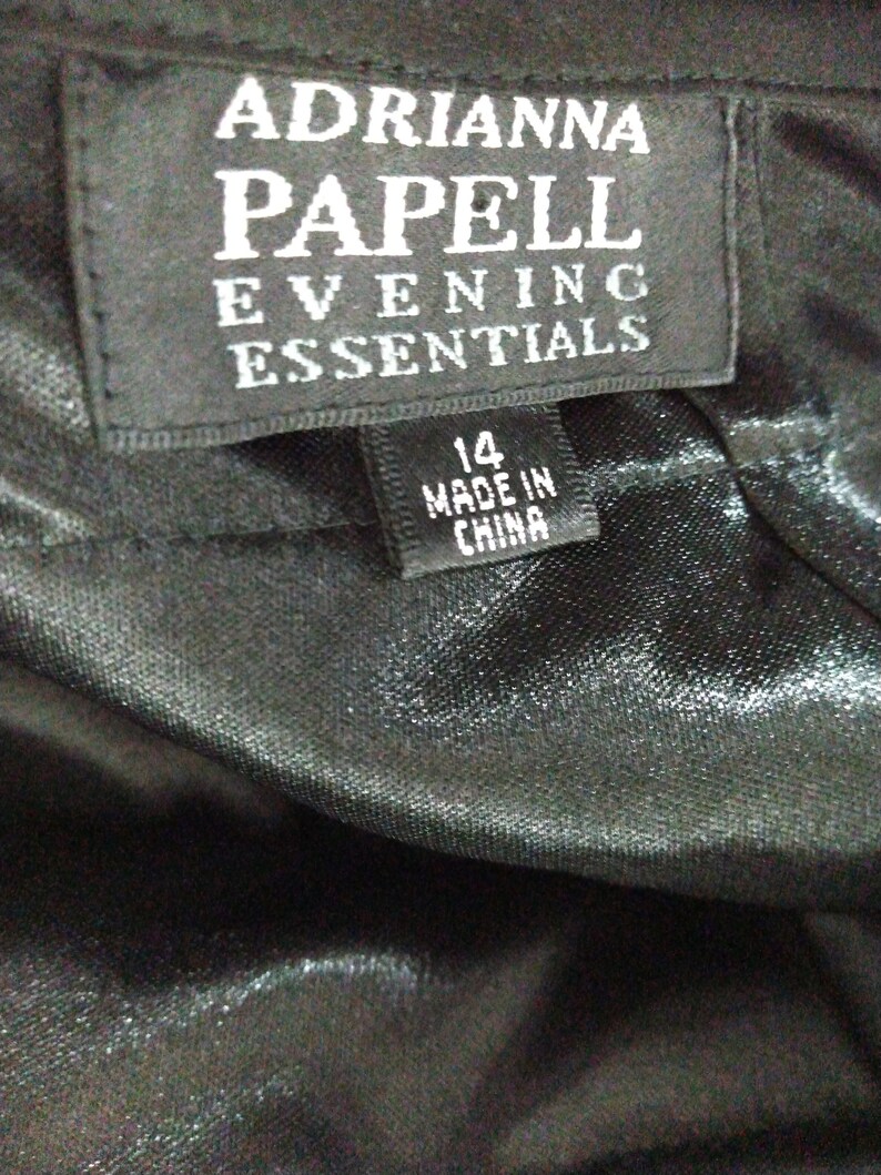 K&ouml;nnte beinhalten: Schwarzes Etikett mit wei&szlig;em Text, der "Adrianna Papell Evening Essentials" lautet, und ein kleineres wei&szlig;es Etikett mit schwarzem Text, der "14 Made in China" lautet.