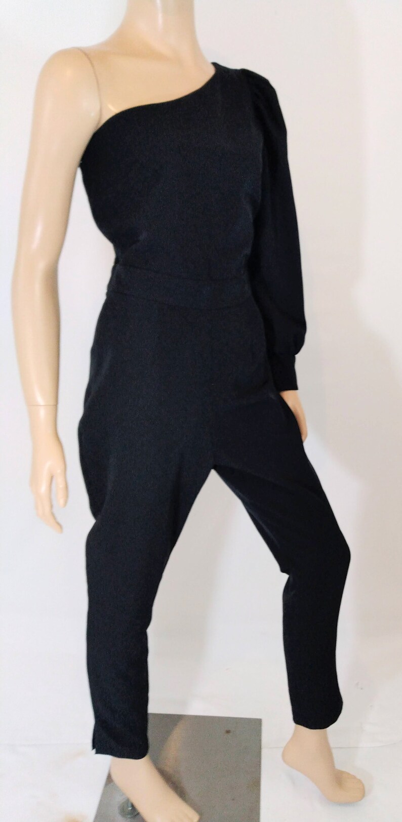 May include: A black one-shoulder jumpsuit with a long sleeve on the right side. The jumpsuit has a fitted bodice and wide-leg pants.