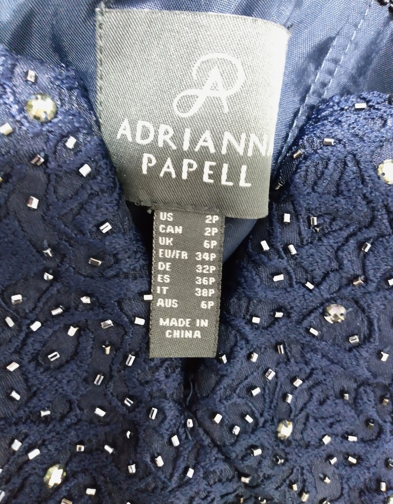 May include: A navy blue dress tag with the brand name "Adrianna Papell" and size information in US, Canadian, UK, EU/FR, German, Spanish, Italian, and Australian sizes. The tag also states "Made in China."