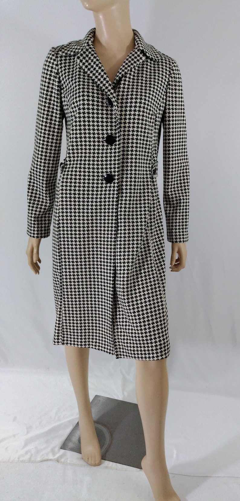 May include: A black and white houndstooth patterned coat with a long sleeve and button closure.