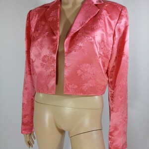 Pu&ograve; includere: Un blazer rosa corto, senza chiusura, con un motivo floreale in rilievo. Presenta maniche lunghe e un classico colletto a revers. Il tessuto satinato conferisce alla giacca un aspetto lucido ed elegante.