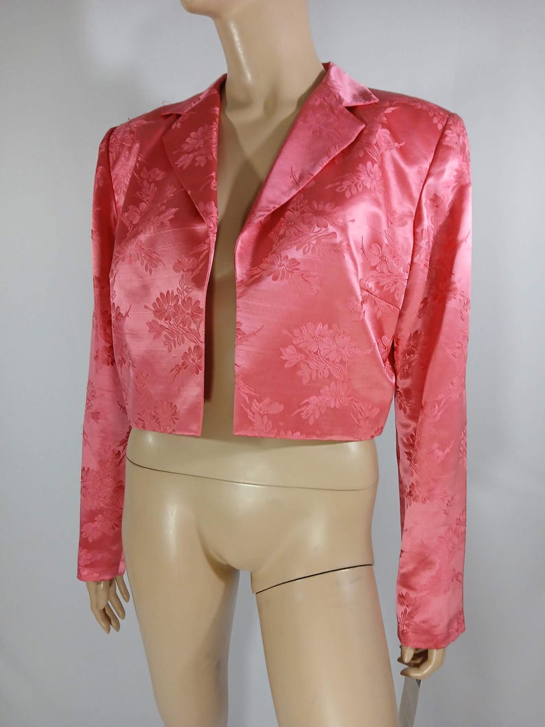 Pu&ograve; includere: Un blazer rosa corto, senza chiusura, con un motivo floreale in rilievo. Presenta maniche lunghe e un classico colletto a revers. Il tessuto satinato conferisce alla giacca un aspetto lucido ed elegante.